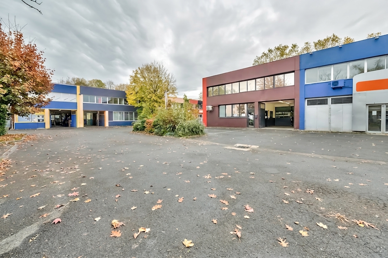 Deux bâtiments modernes et colorés se dressent de part et d'autre d'un terrain asphalté presque désert, parsemé de feuilles d'automne, avec quelques arbres en arrière-plan sous un ciel nuageux.