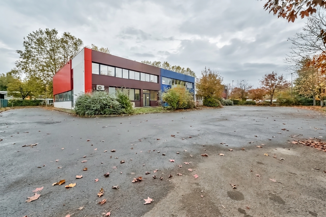 Un immeuble de bureaux de deux étages, recouvert de panneaux rouges et marron, entouré d'arbres et d'un parking asphalté désert, par une journée nuageuse.
