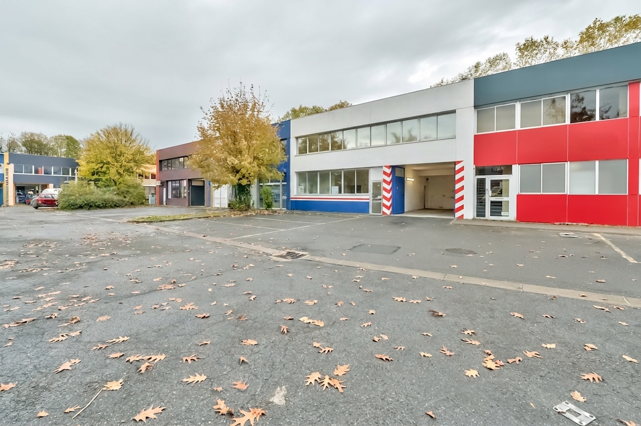 Un immeuble commercial aux panneaux rouges, bleus et blancs se dresse à côté d’un parking vide jonché de feuilles d’automne tombées, sous un ciel nuageux.