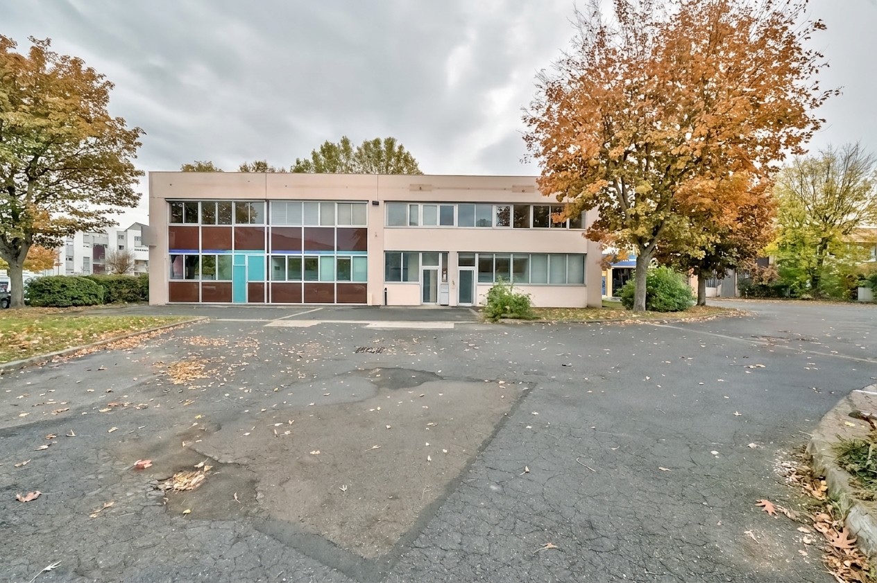 Immeuble commercial de deux étages doté de grandes fenêtres, d'un toit plat et d'un parking vide devant, entouré d'arbres aux couleurs automnales sous un ciel nuageux.