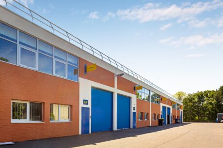 Une rangée d'unités industrielles modernes avec des briques orange et des murs blancs, des portes bleues et des enseignes, le tout sous un ciel clair.