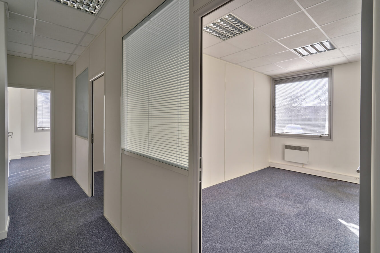 Un intérieur de bureau moderne avec des pièces vides, des sols recouverts de moquette, des murs blancs, de grandes fenêtres et des plafonniers fluorescents.