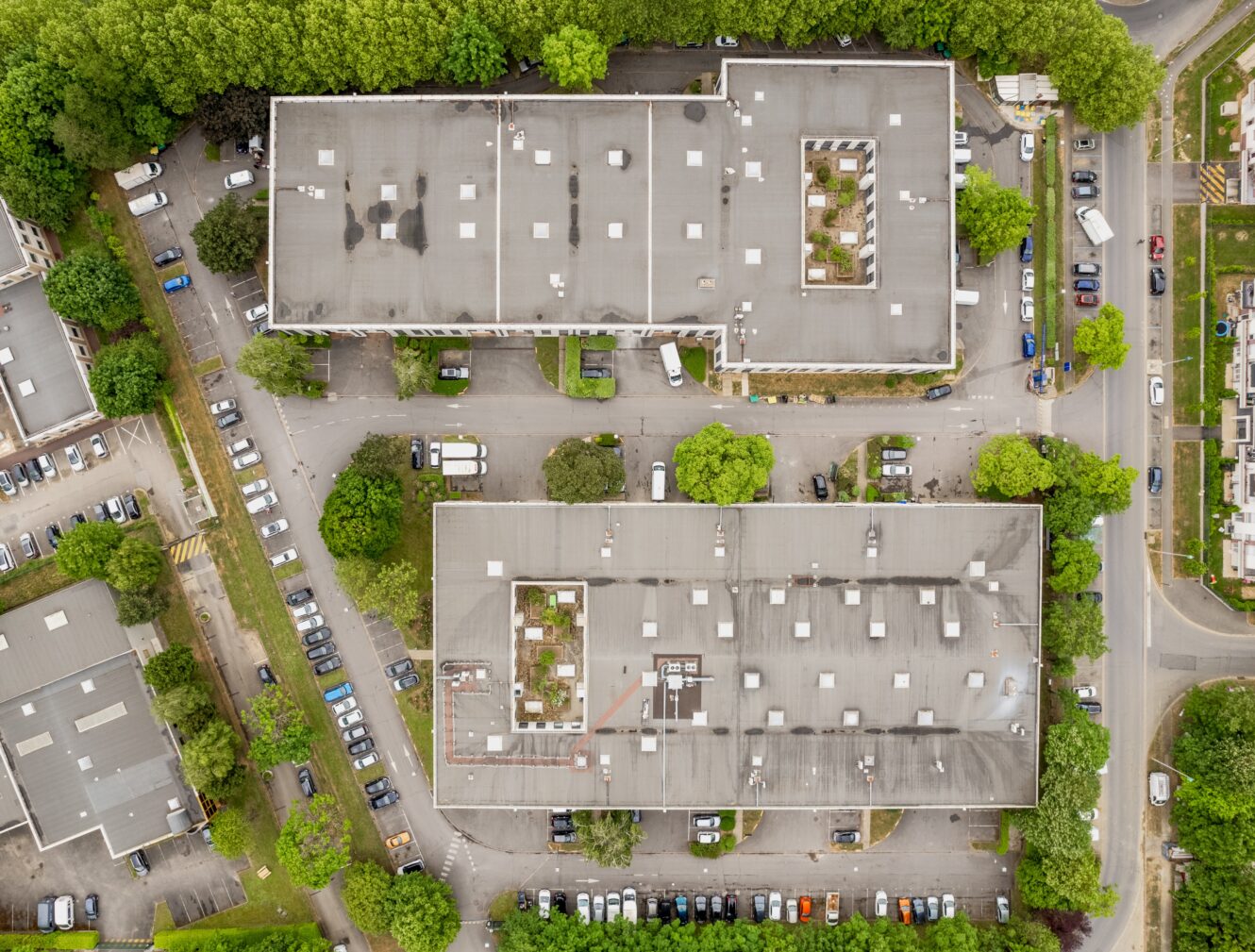 Vue aérienne de deux grands bâtiments rectangulaires entourés d'arbres, de voitures garées et de routes dans une zone urbaine.