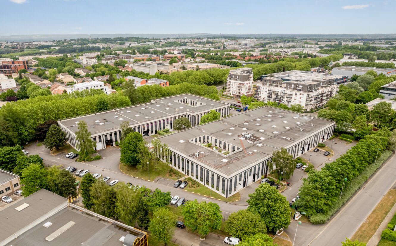 Vue aérienne d'un parc d'activités avec plusieurs immeubles de bureaux rectangulaires entourés d'arbres, de voitures garées et de zones résidentielles et commerciales à proximité.