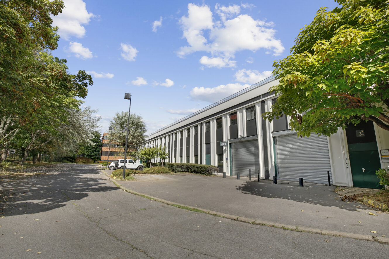 Une large rue passe devant un bâtiment industriel blanc avec de grandes fenêtres et des portes de garage, entouré d'arbres sous un ciel partiellement nuageux.