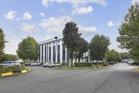 Un immeuble de bureaux moderne de trois étages avec des panneaux rayés noirs et blancs, entouré d'arbres et de voitures garées, sous un ciel partiellement nuageux.