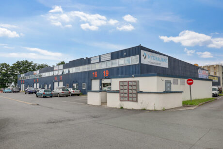 Un bâtiment commercial avec des unités numérotées et des voitures garées devant, avec l'enseigne Hydralians sur le côté, sous un ciel partiellement nuageux.