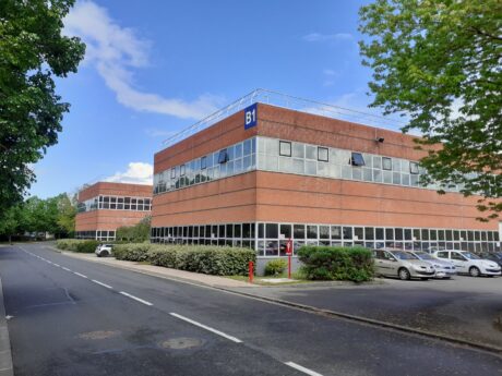 Un immeuble de bureaux moderne en briques étiqueté B1 avec de grandes fenêtres, un parking avec des voitures, des arbres et un ciel dégagé en arrière-plan.