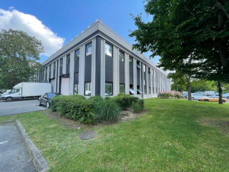 Un immeuble de bureaux moderne de deux étages avec de grandes fenêtres, entouré d'herbe, d'arbres et de véhicules garés sous un ciel bleu.