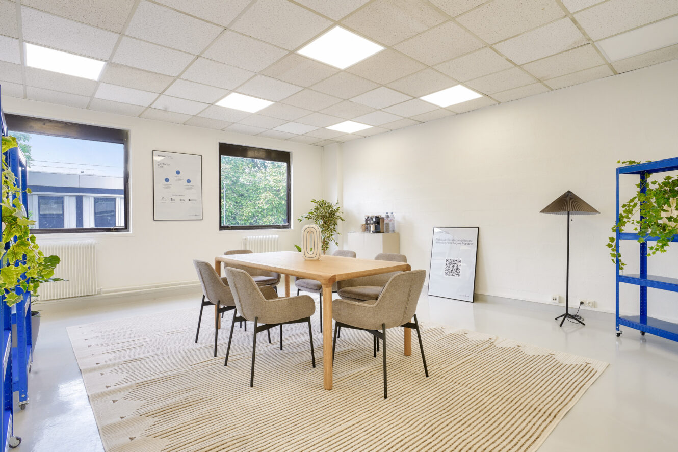 Une salle de réunion de bureau moderne avec une table en bois, quatre chaises, des plantes en pot, un tapis et de grandes fenêtres laissant entrer la lumière naturelle.
