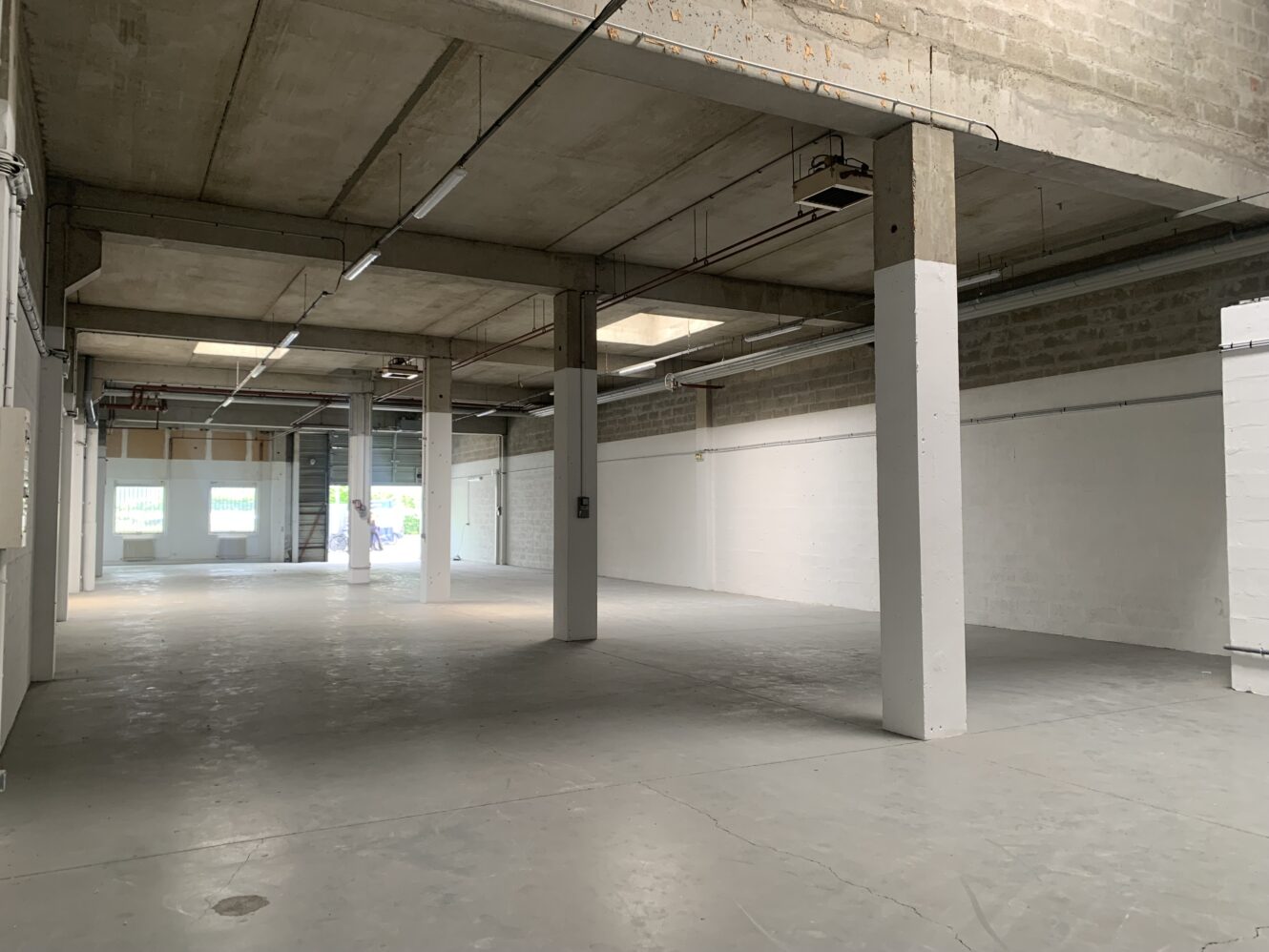 Spacieux entrepôt industriel vide avec des sols en béton, des colonnes, des plafonds apparents et des éclairages fluorescents. La lumière naturelle pénètre par les fenêtres et les portes ouvertes à l'extrémité.