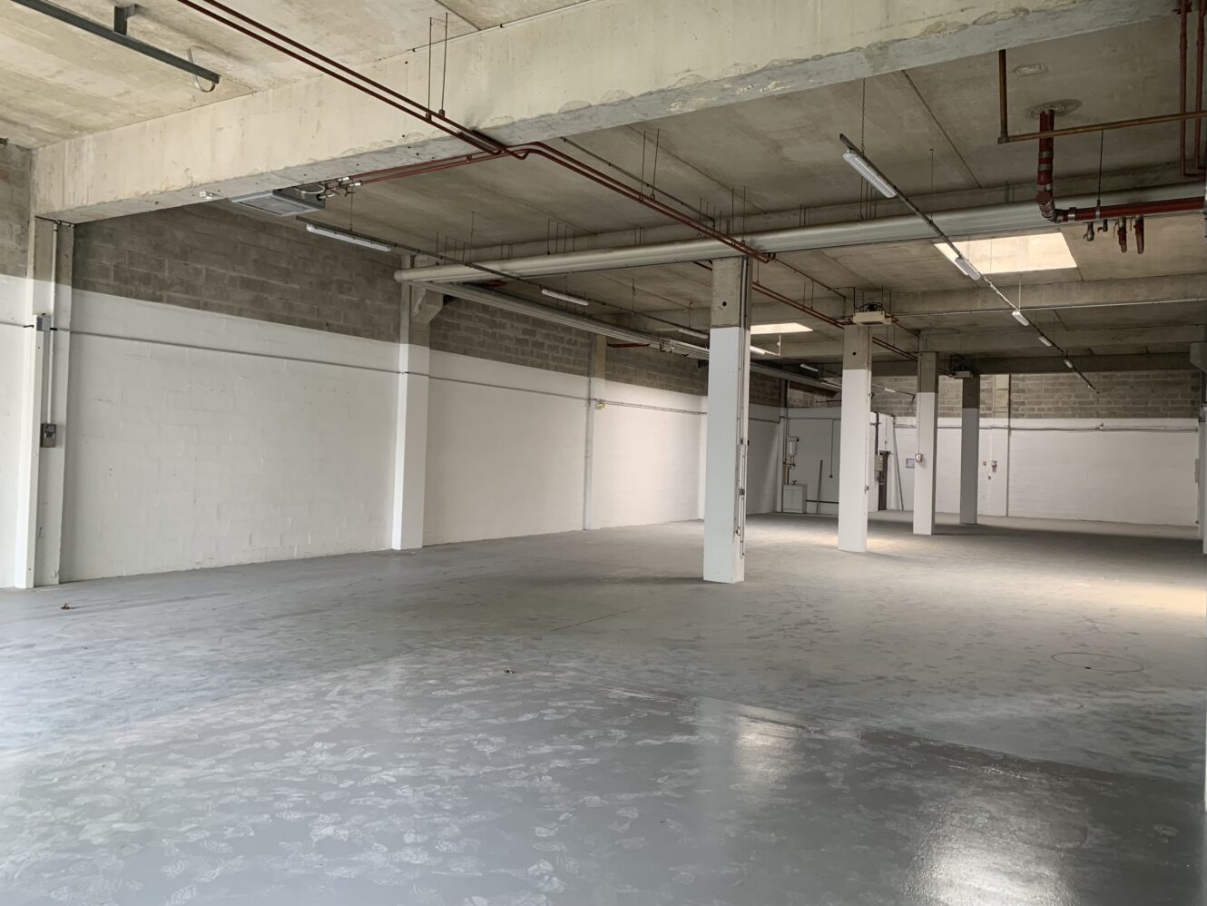 Entrepôt industriel vide avec sol en béton, tuyaux de plafond apparents et murs en briques peintes en blanc sous éclairage naturel et artificiel.