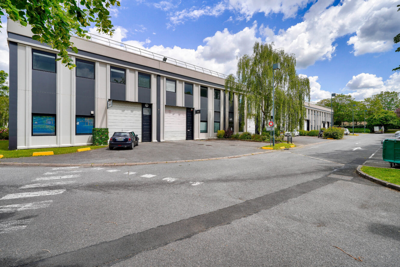 Bâtiment commercial moderne de deux étages avec de grandes fenêtres, des places de parking et quelques voitures garées devant, entouré d'arbres et de verdure sous un ciel partiellement nuageux.