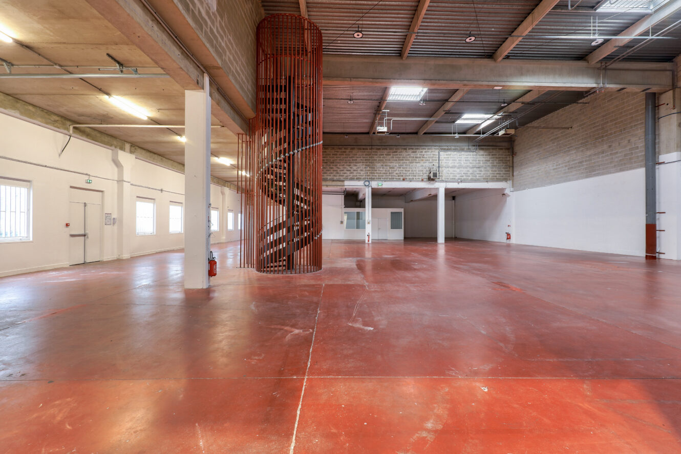 Spacieux entrepôt vide avec un sol en béton rouge, un haut plafond, des poutres apparentes et un escalier en colimaçon au centre. Des murs blancs et des fenêtres bordent le côté gauche.