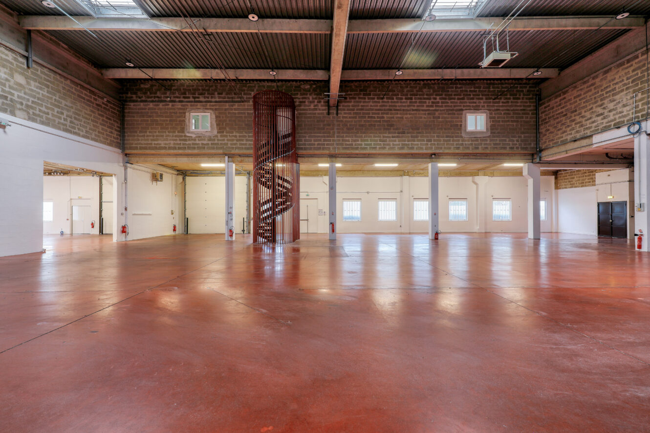 Grand entrepôt industriel vide avec un sol en béton rouge, un haut plafond, des murs en briques apparentes et un escalier central en colimaçon. Plusieurs fenêtres et portes bordent le mur du fond.