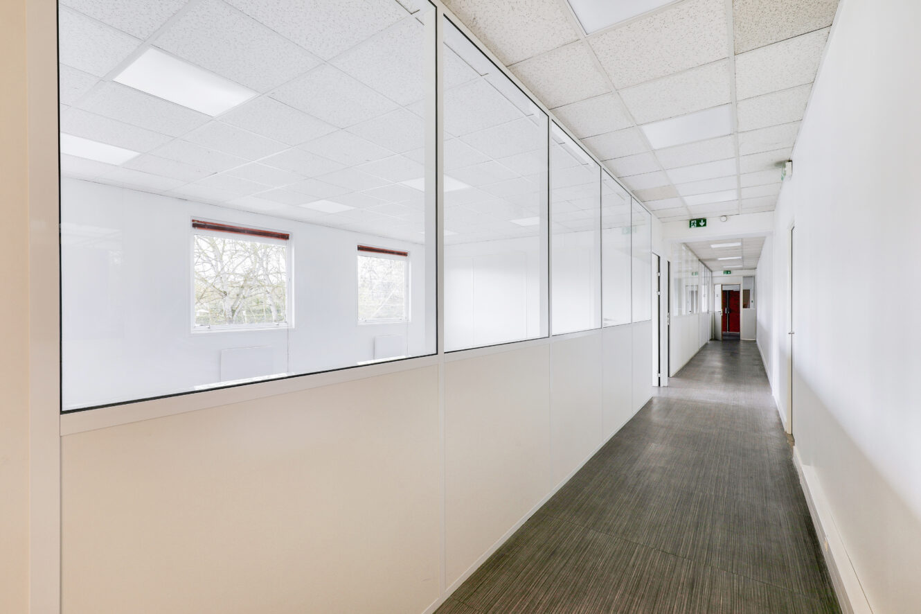 Un long couloir de bureau vide avec des pièces vitrées d'un côté, des murs blancs et un plafond avec des lumières fluorescentes.