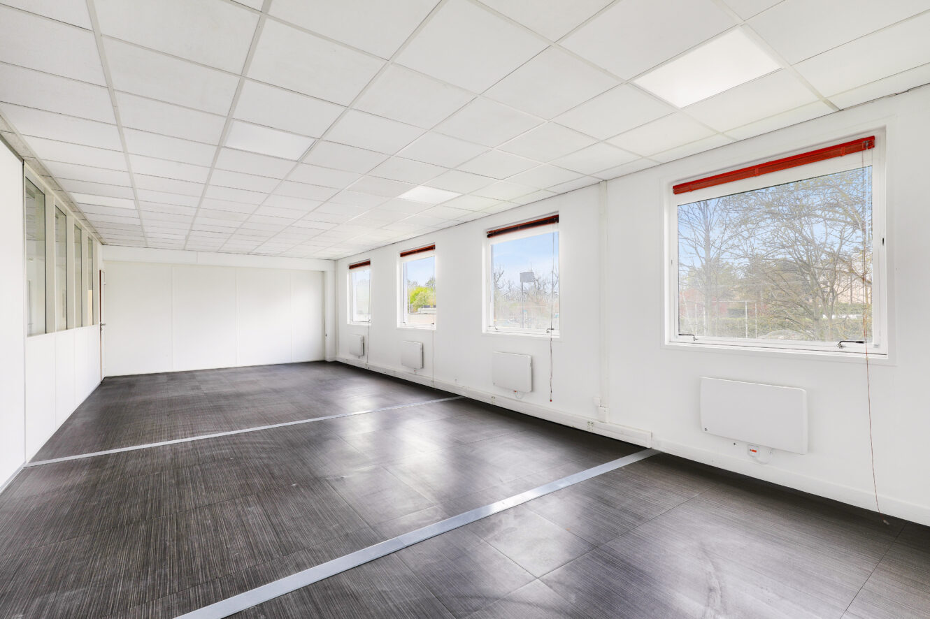 Bureau vide aux murs blancs, aux grandes fenêtres, au sol carrelé noir et au plafond quadrillé de lampes fluorescentes.