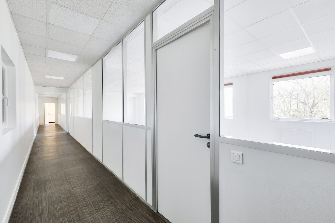 Un couloir avec des murs blancs, des cloisons de bureau en verre, des portes blanches fermées et un sol en bois dans un immeuble de bureaux moderne.
