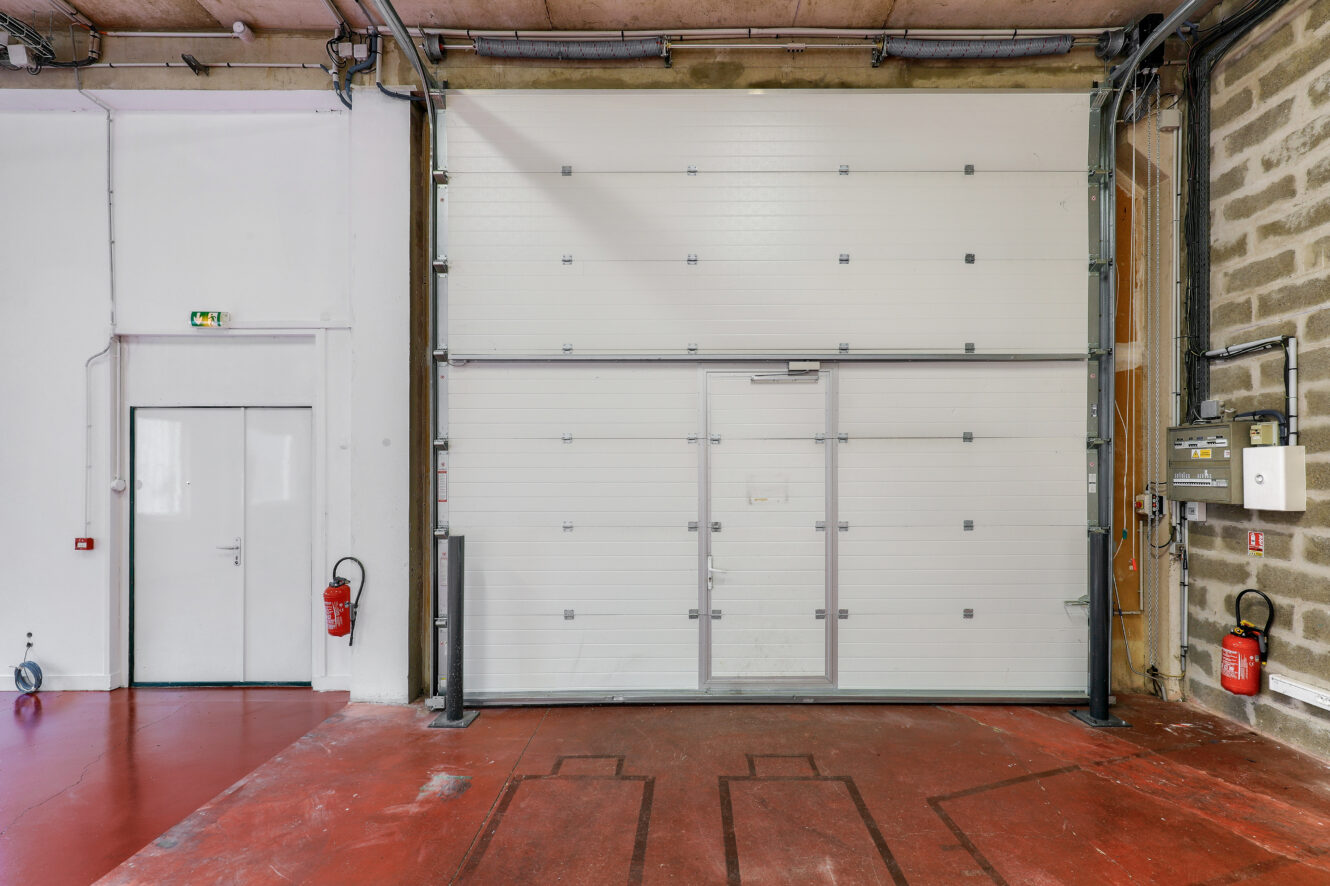 Grande porte de garage industrielle à côté d'une porte piétonne blanche, d'extincteurs et de panneaux électriques sur un sol rouge et en béton.