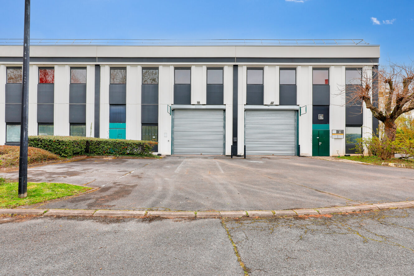 Un entrepôt commercial avec deux grandes portes de chargement fermées, des portes d'entrée vertes et un parking vide à l'avant.