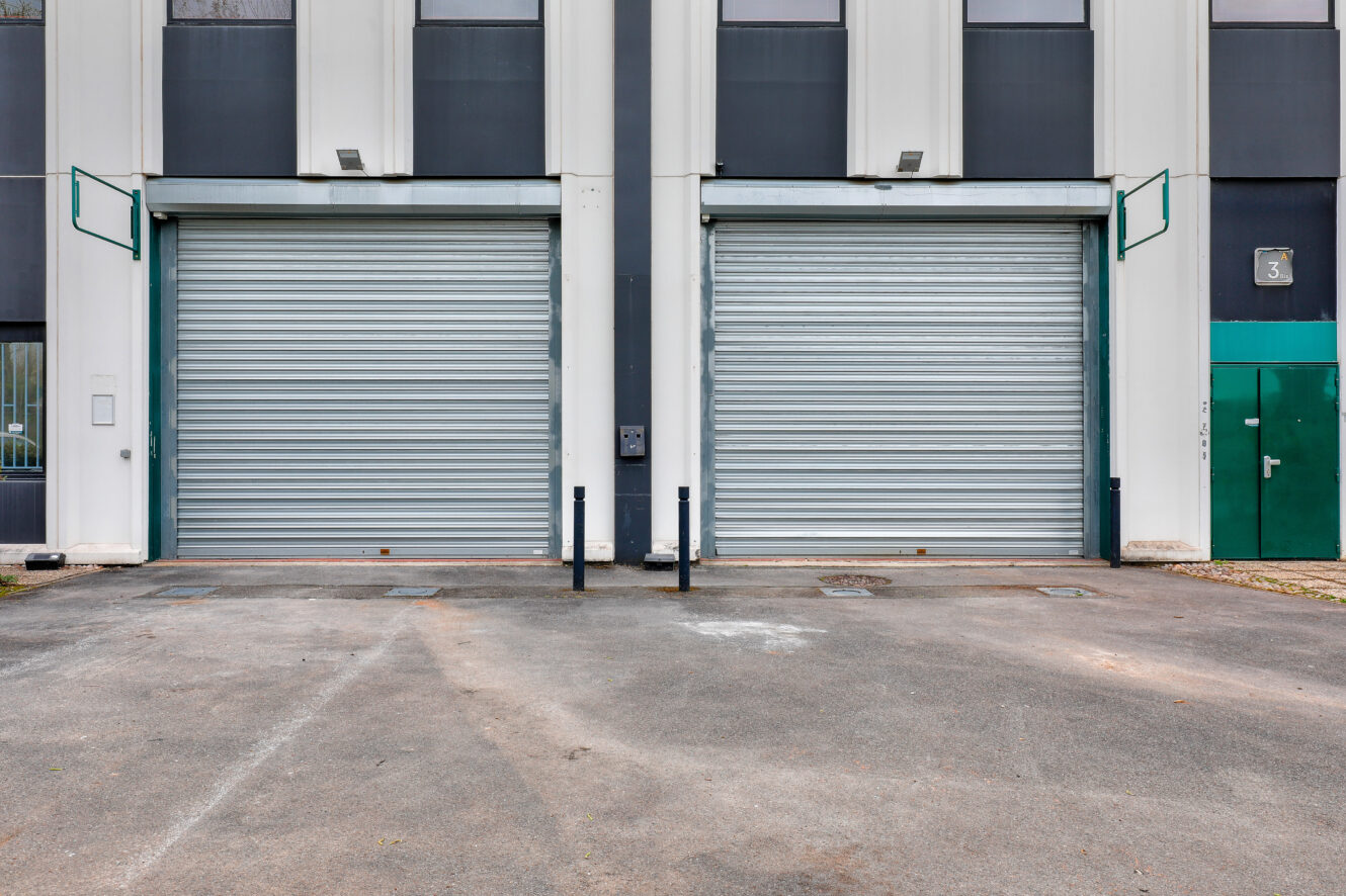 Deux grandes portes métalliques à enroulement, grises et fermées, sur un bâtiment industriel, avec une petite porte verte à droite et des bornes devant les entrées.
