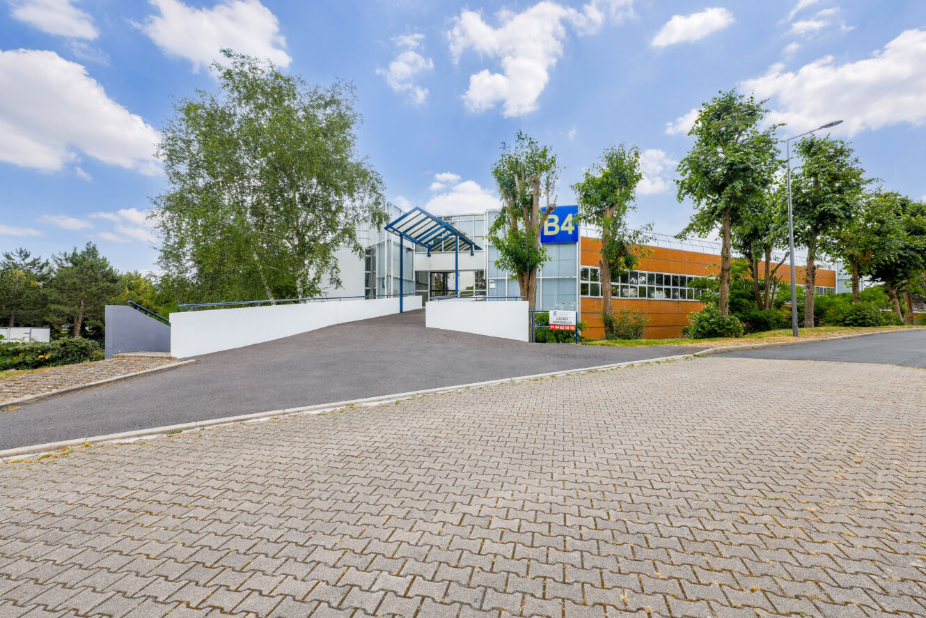 Immeuble de bureaux moderne avec entrée vitrée, étiqueté B4, entouré d'arbres, d'une allée pavée et d'un trottoir, sous un ciel partiellement nuageux.