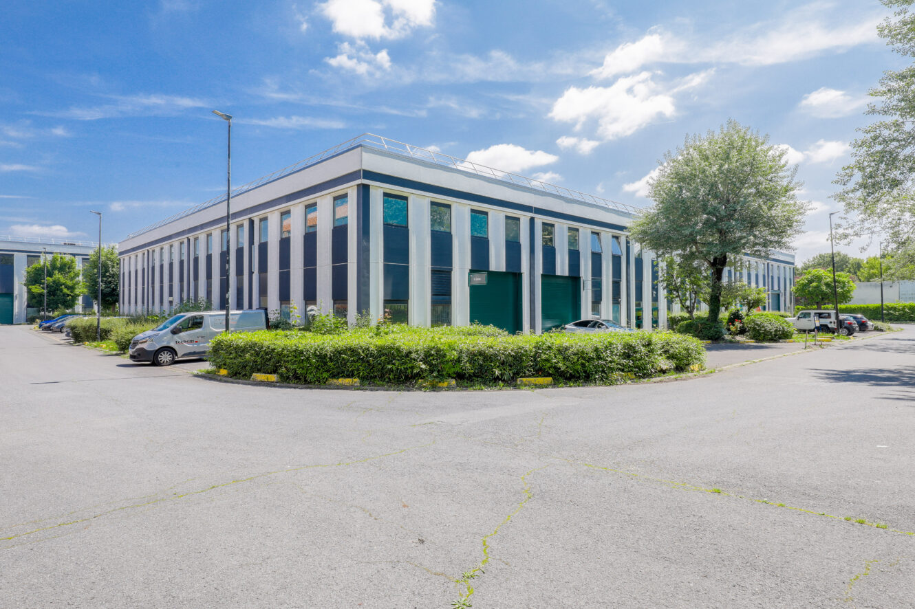 Immeuble de bureaux moderne de deux étages avec de grandes fenêtres, entouré de verdure et de voitures garées sous un ciel partiellement nuageux.