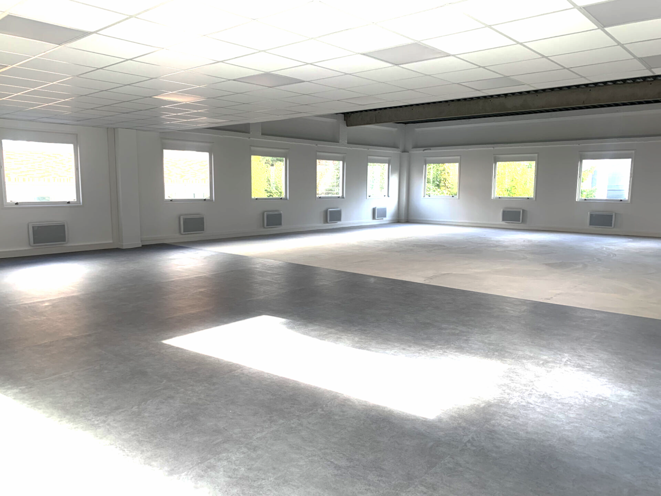 Pièce spacieuse et vide avec de grandes fenêtres, des murs blancs et un plafond en grille ; la lumière du soleil pénètre à l'intérieur et éclaire une partie du sol gris.