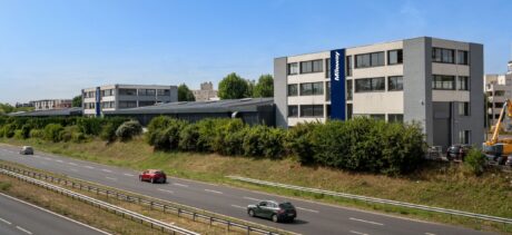 Trois voitures roulent sur une autoroute à chaussées séparées à côté d'immeubles de bureaux modernes, dont l'un affiche un panneau indiquant Midway. Des arbustes bordent la route et des arbres se trouvent à l'arrière-plan.