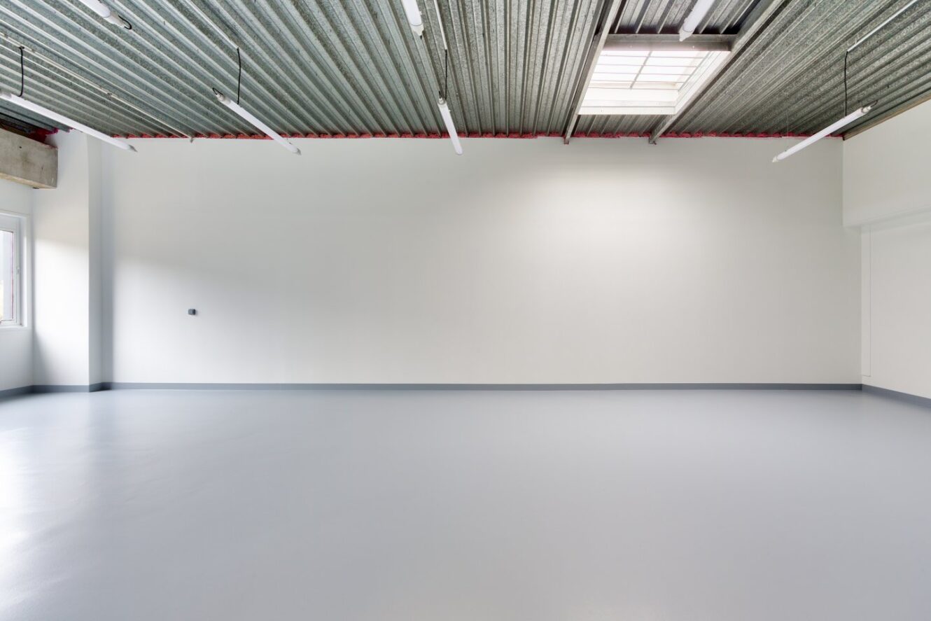 Pièce vide de style industriel avec des murs blancs, un sol gris lisse, des lampes fluorescentes et un plafond en tôle ondulée avec un puits de lumière.