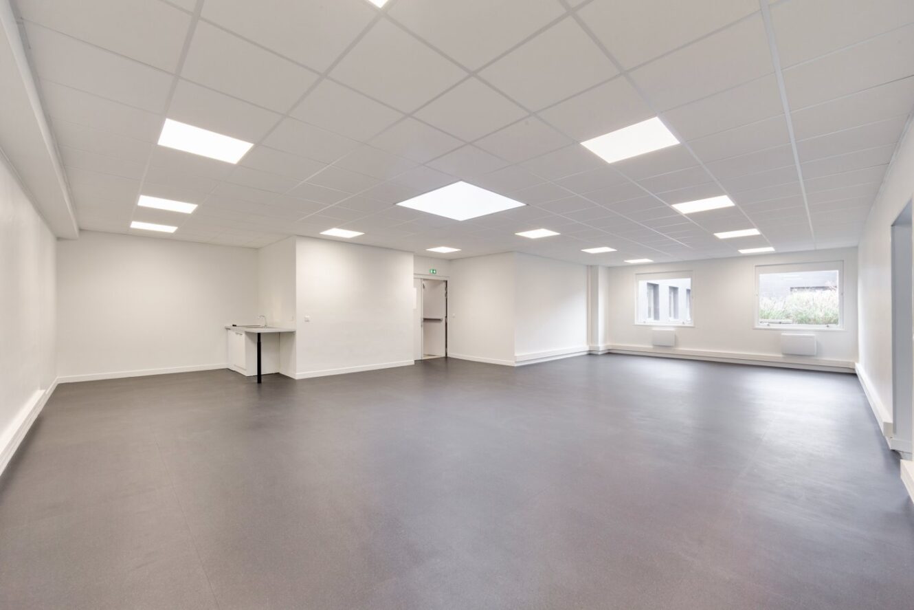 Pièce spacieuse et vide avec des murs blancs, des plafonniers, un sol sombre, deux fenêtres et une porte au fond.