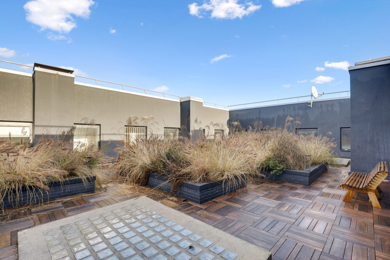Un toit-terrasse avec une terrasse en bois, des jardinières remplies d'herbes hautes, un banc et des murs de bâtiments modernes gris sous un ciel bleu avec des nuages.
