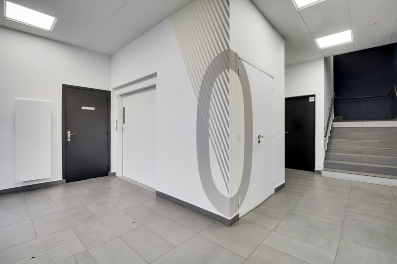 Un couloir moderne avec un carrelage gris, des murs blancs, un ascenseur, deux portes noires, un escalier et un art mural géométrique avec un grand motif circulaire.