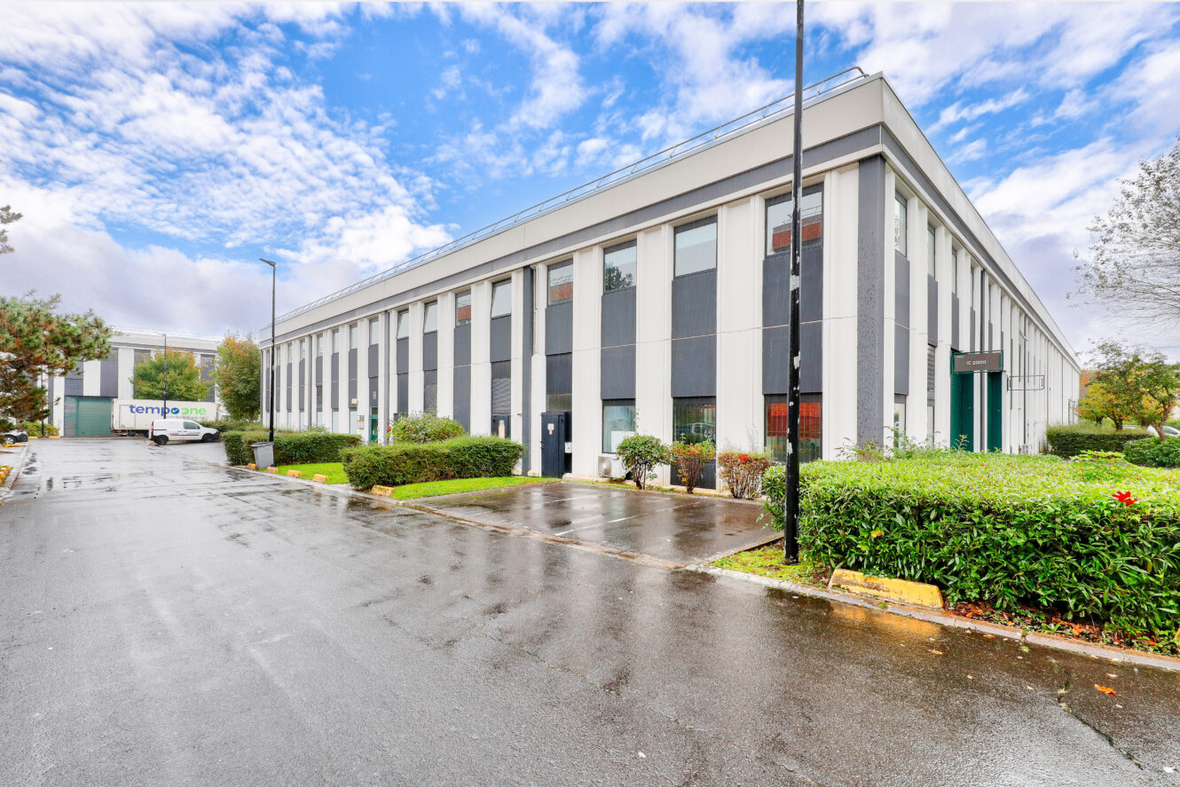 Un immeuble de bureaux moderne de deux étages avec de grandes fenêtres, entouré de verdure, dans une rue humide sous un ciel partiellement nuageux.