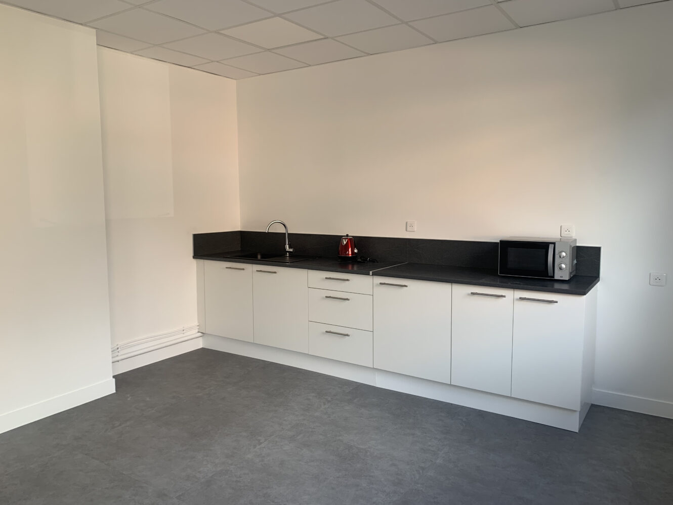 Kitchenette moderne avec armoires blanches, comptoir noir, évier, bouilloire rouge et micro-ondes, sur un mur blanc uni et un sol gris.