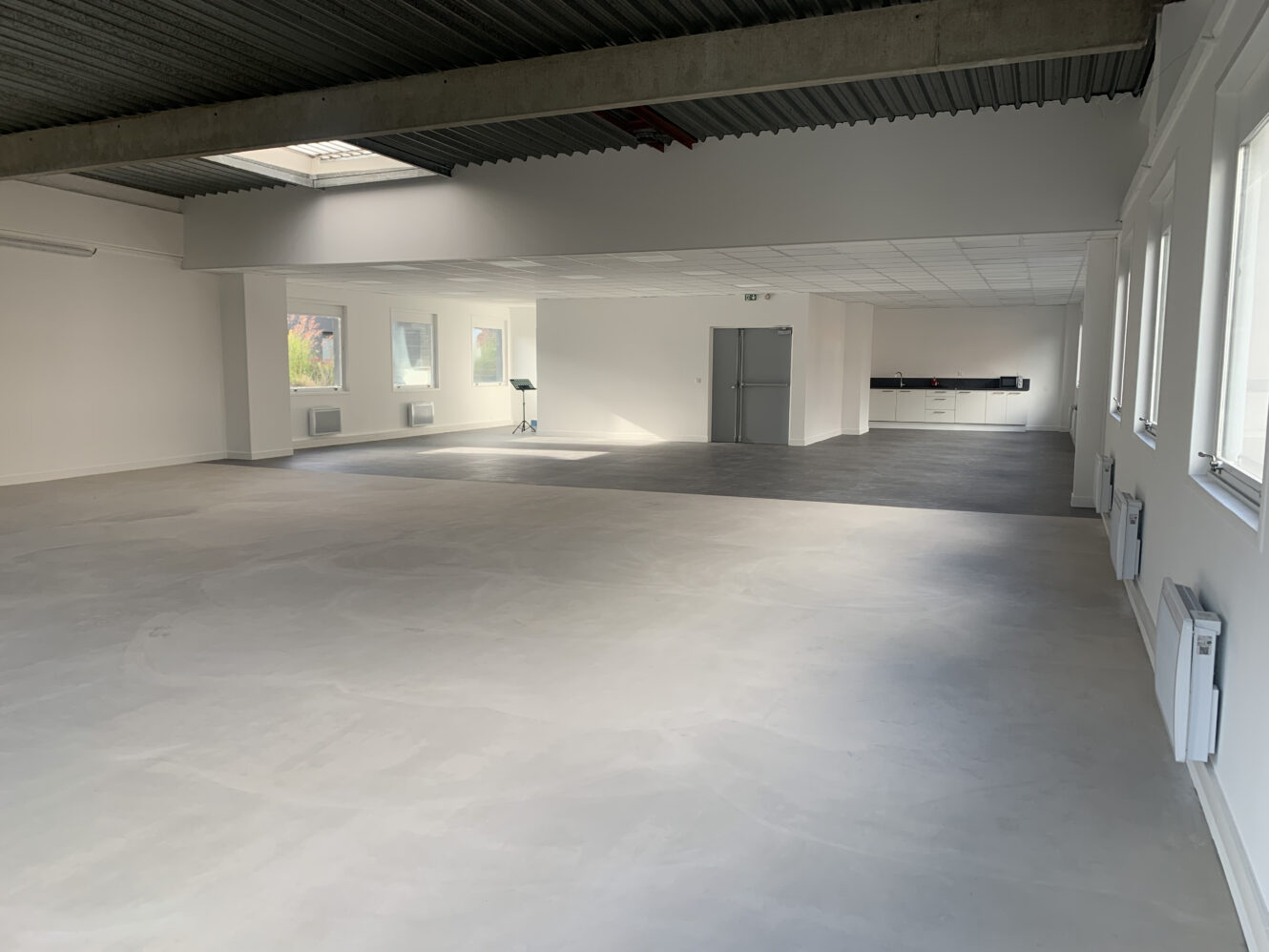 Une pièce spacieuse et vide avec de grandes fenêtres, des murs blancs et un sol gris. Il y a quelques chaises et tables au fond et la lumière naturelle entre par les fenêtres.