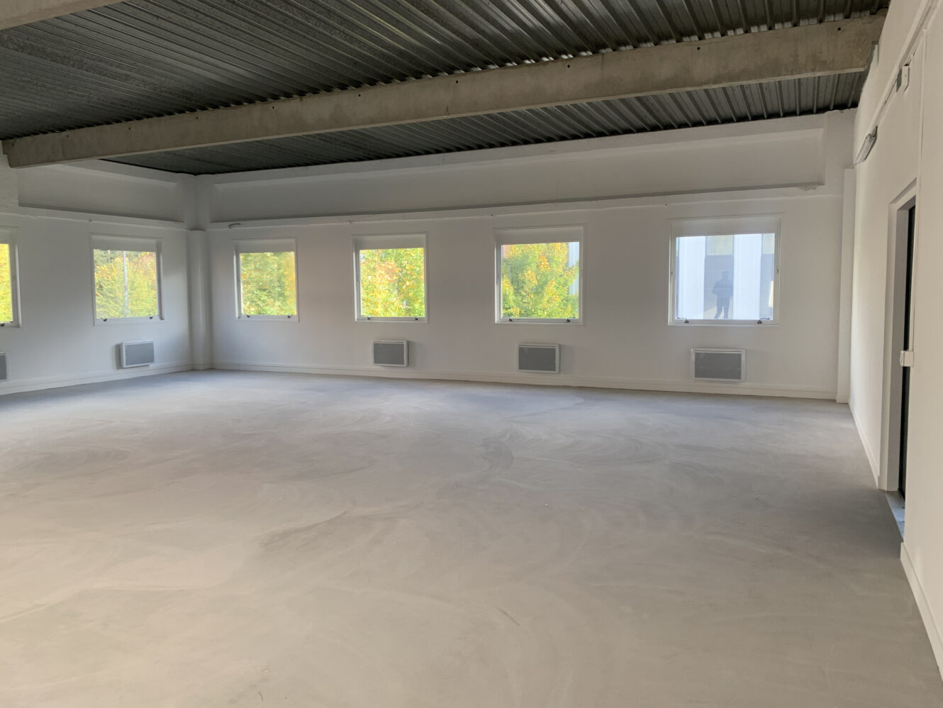 Pièce vide, non meublée, avec un sol en béton, des murs blancs, de grandes fenêtres laissant entrer la lumière naturelle et un plafond en tôle ondulée.