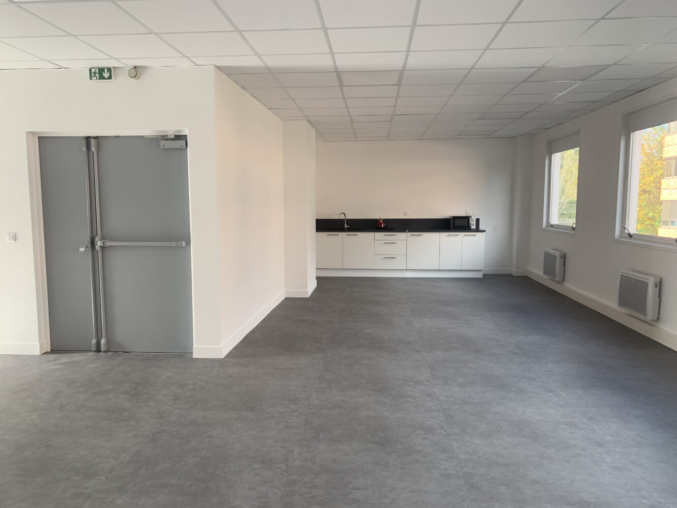 Une pièce spacieuse et vide avec un sol gris, des murs blancs, de grandes fenêtres, une double porte grise et une petite kitchenette avec un évier et un micro-ondes dans le coin arrière.