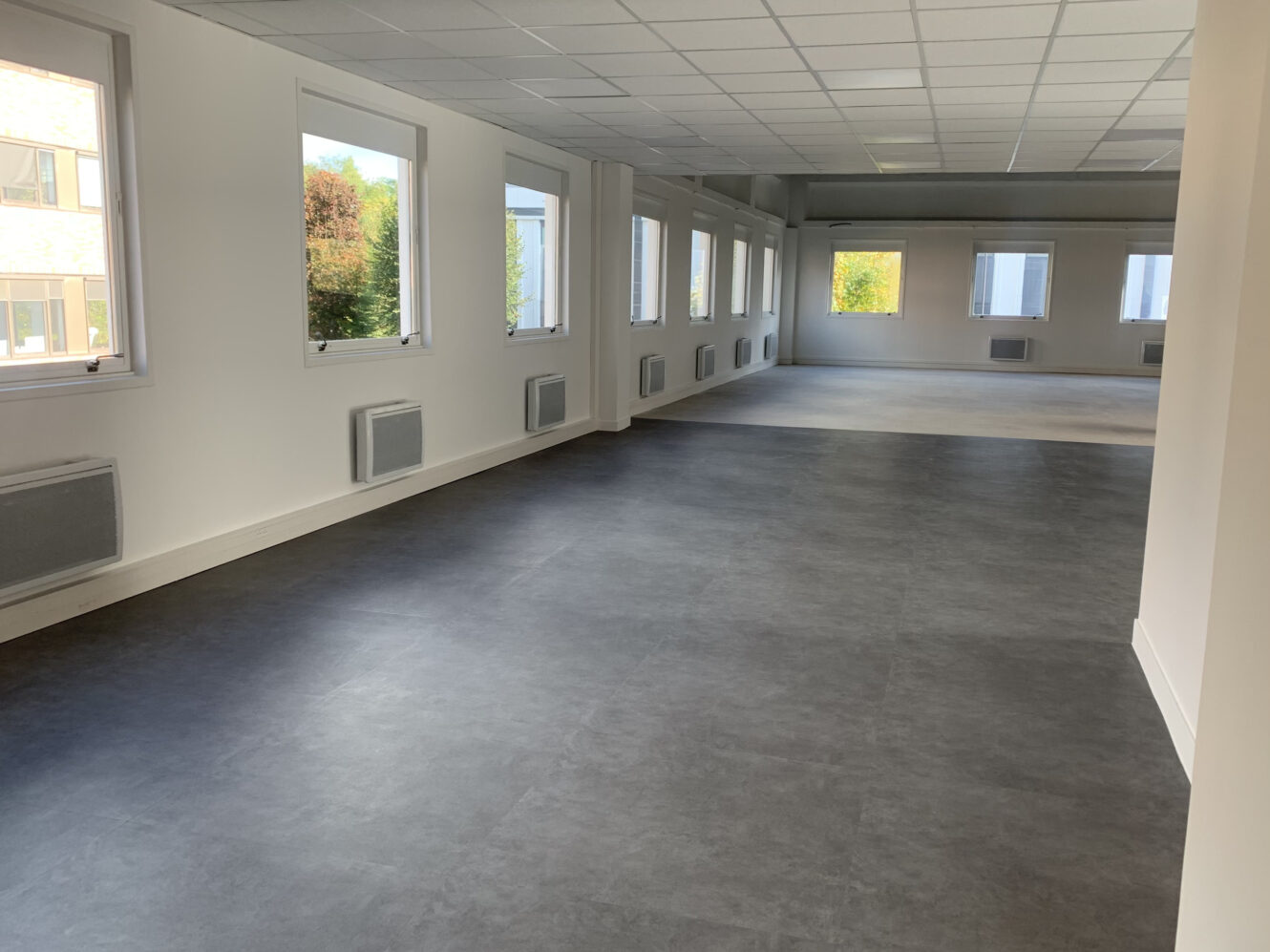 Bureau vide avec de grandes fenêtres, des murs blancs, un sol gris et des radiateurs muraux, la lumière naturelle entrant par les fenêtres.