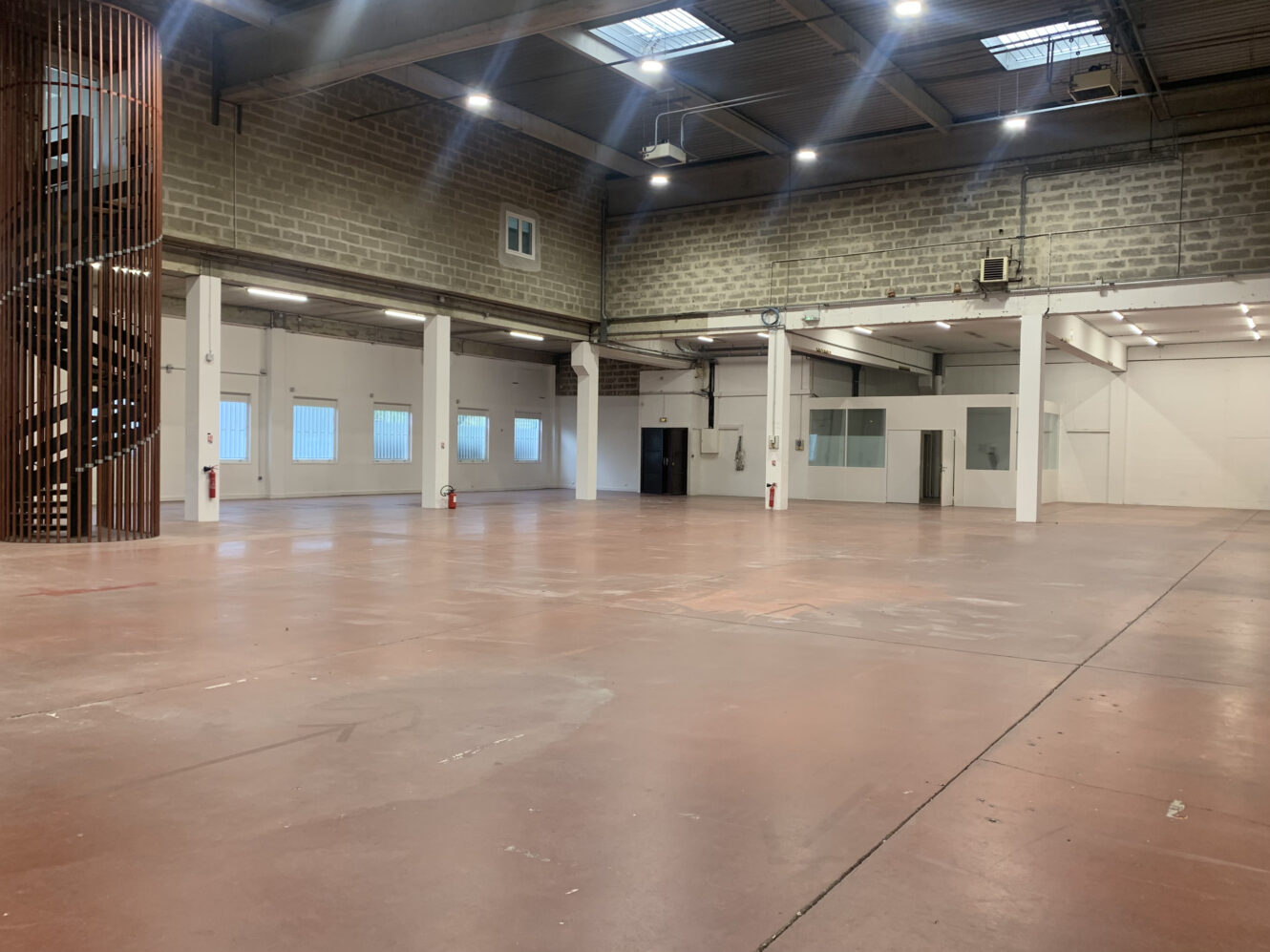 Grand entrepôt vide avec de hauts plafonds, des murs en briques apparentes, des fenêtres sur un côté, des extincteurs et un escalier en colimaçon sur la gauche.