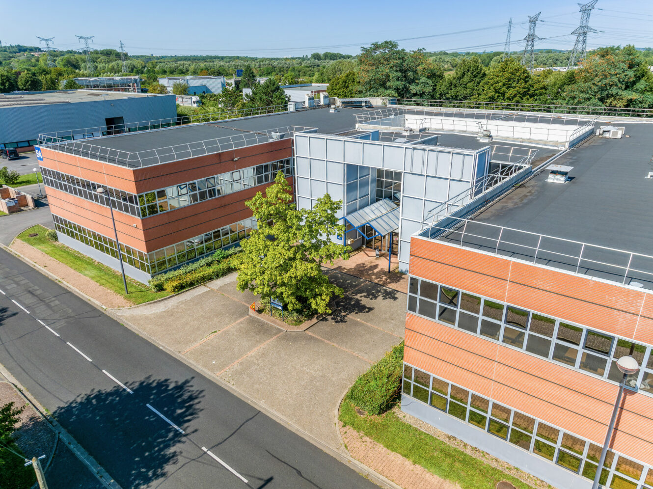 Vue aérienne d'un complexe d'immeubles de bureaux modernes aux façades de briques rouges et de verre, entouré d'arbres et de routes, sous un ciel dégagé.