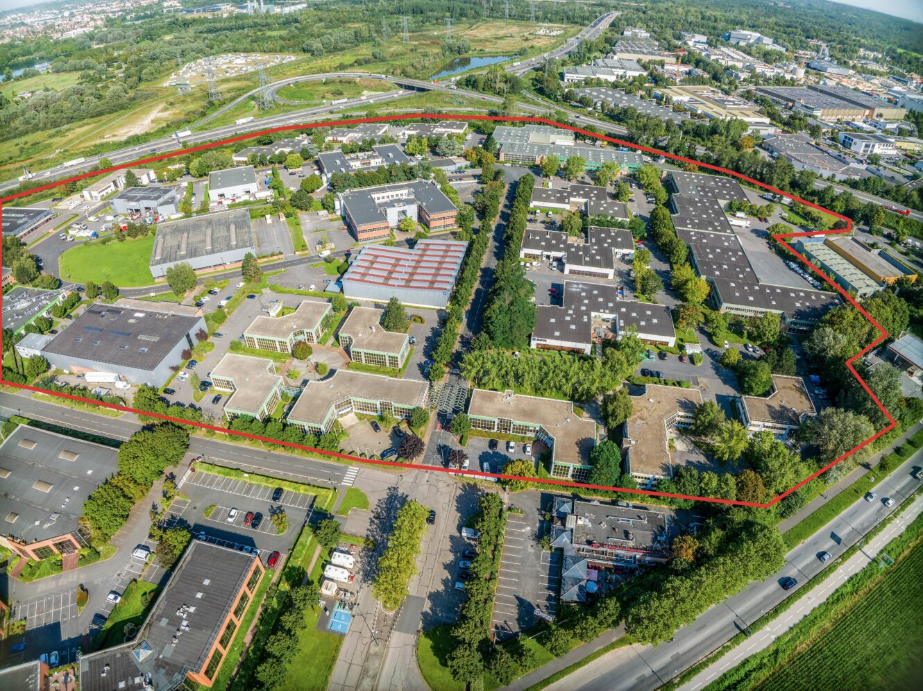 Vue aérienne d'un complexe industriel et de bureaux, délimité par un cadre rouge, entouré de verdure, de routes et d'autres bâtiments.