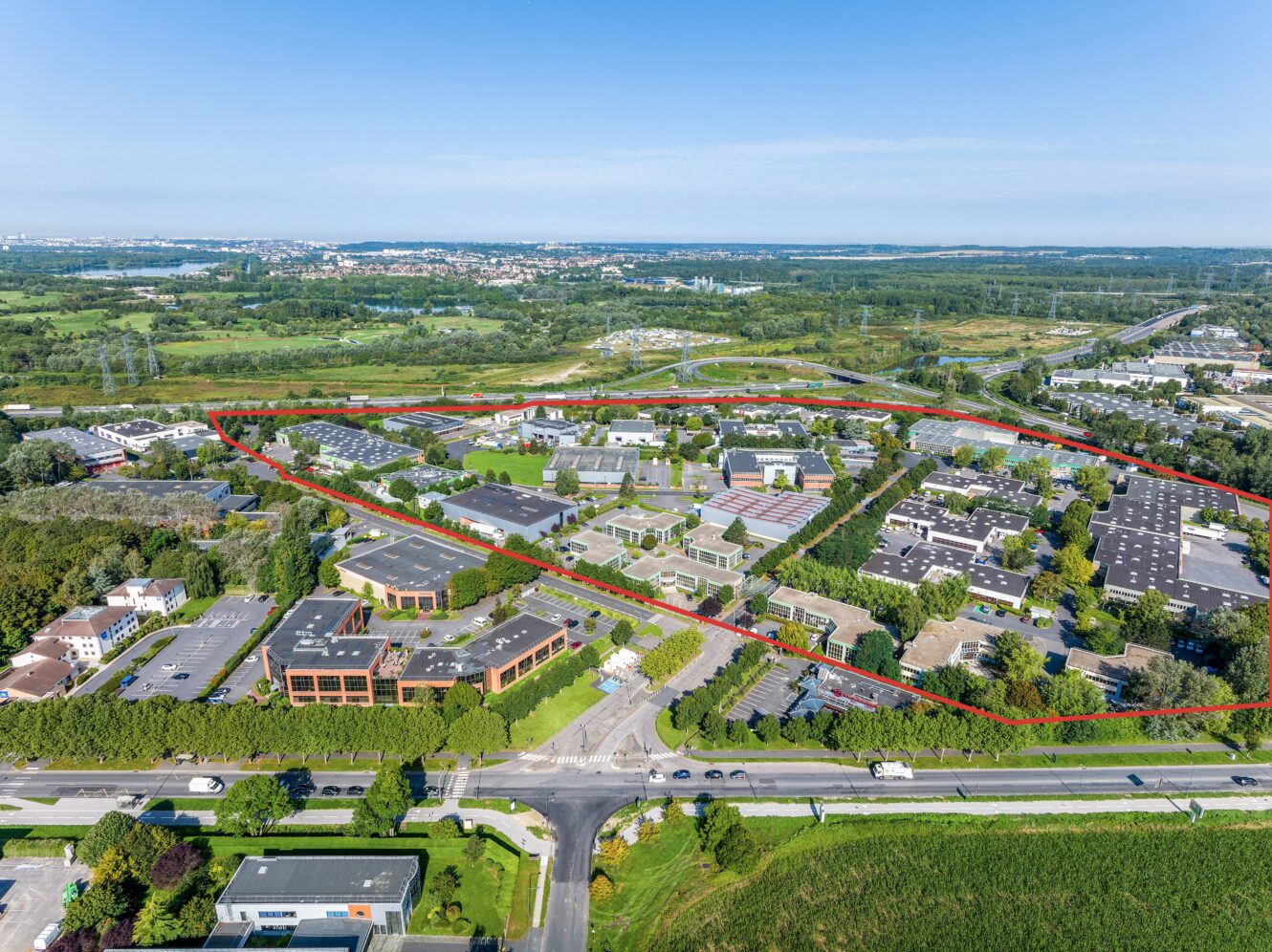 Vue aérienne d'un parc commercial ou industriel avec de nombreux bâtiments, des arbres et des parkings délimités par un cadre rouge, entourés de routes et d'espaces verts.