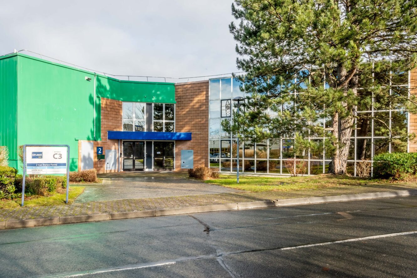 Immeuble de bureaux moderne avec de grandes fenêtres, une façade verte et brune et un auvent d'entrée bleu, entouré d'arbres et d'un trottoir humide devant.