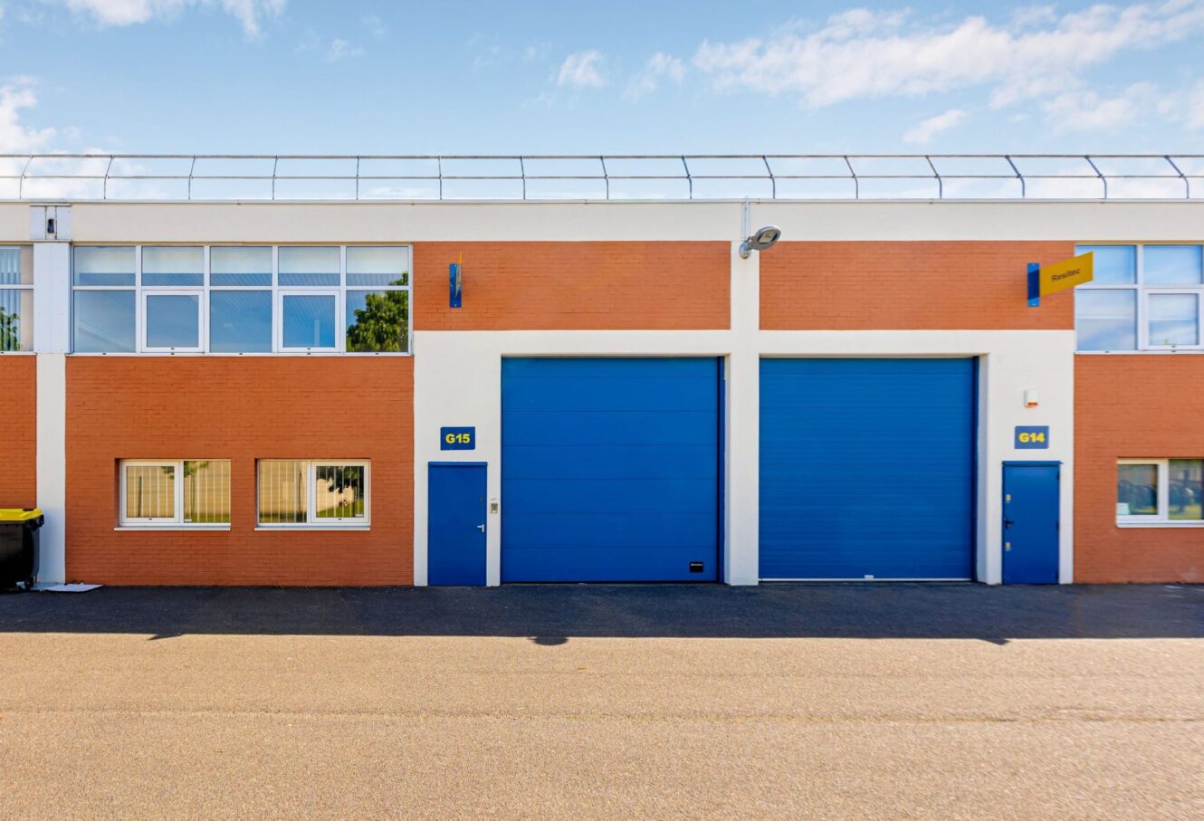 Un bâtiment industriel moderne avec une façade en briques orange, deux grandes portes de garage bleues, plusieurs fenêtres et portes d'entrée, sous un ciel bleu.