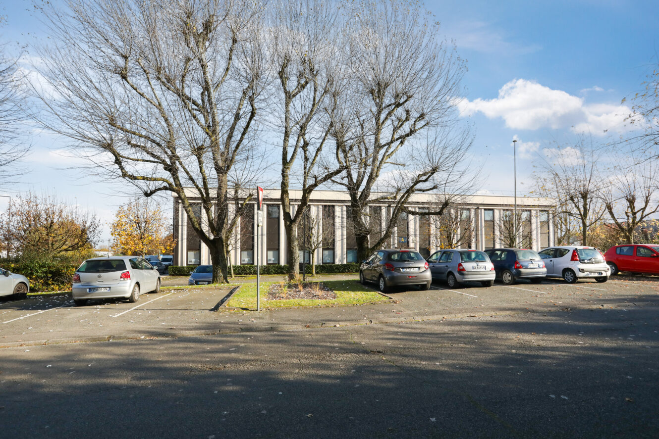 Une rangée de voitures garées dans un parking avec des arbres sans feuilles devant un bâtiment moderne par temps clair.