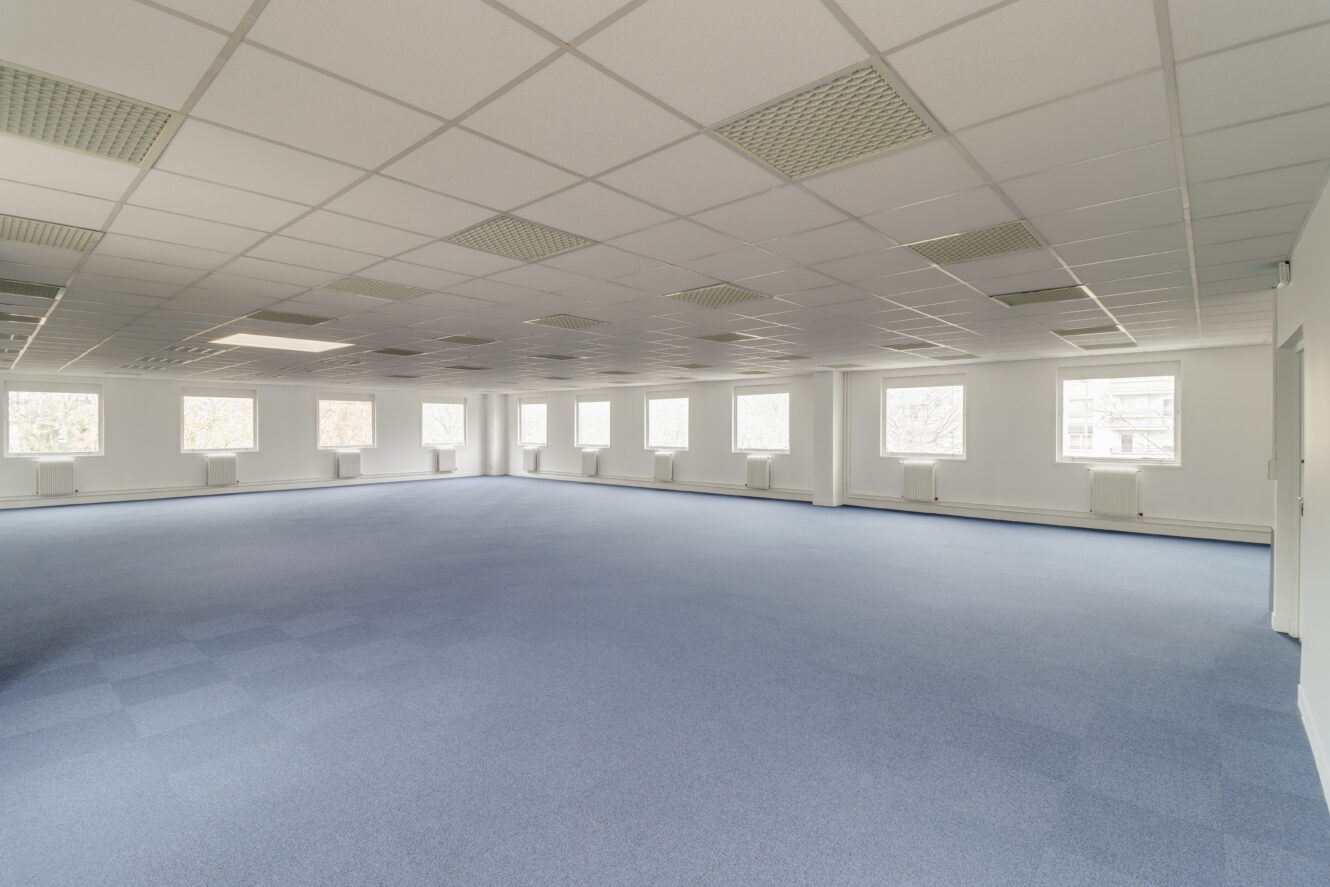 Grand espace de bureau vide avec une moquette bleue, des murs blancs et de nombreuses fenêtres laissant entrer la lumière naturelle.