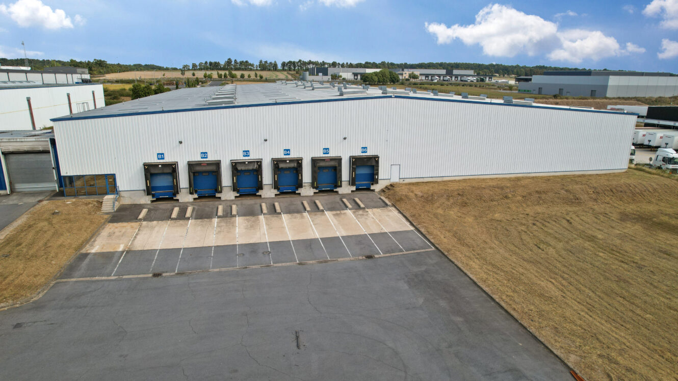 Un grand entrepôt avec six quais de chargement et un parking vide, entouré d'herbe et d'autres bâtiments industriels en arrière-plan.