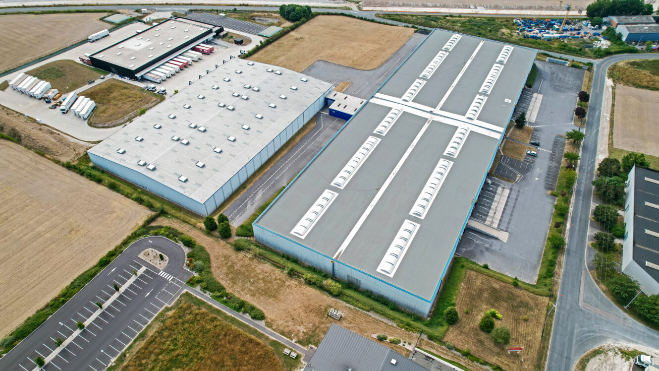Vue aérienne de deux grands entrepôts entourés de parkings, de routes et de champs dans une zone industrielle.