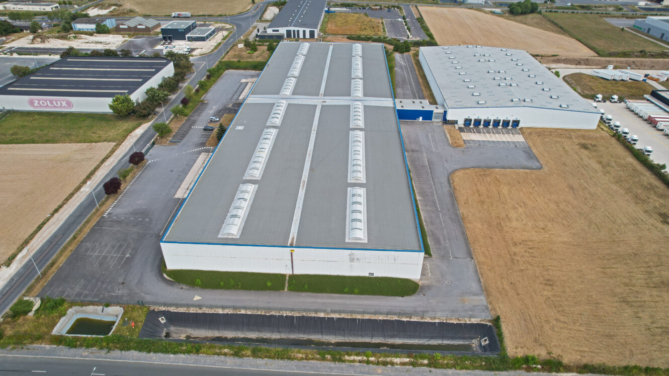 Vue aérienne d'un grand entrepôt industriel au toit gris, entouré de parkings et de champs, dans une zone commerciale.